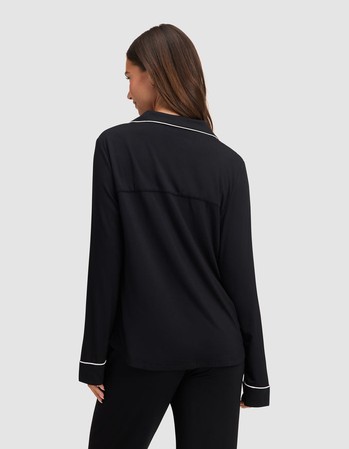 A woman with long brown hair faces away, wearing the HIDE Women's Bamboo Stretch-Knit Long Sleeve Pajama Top in black with white piping, against a plain light gray background. |Color:Black