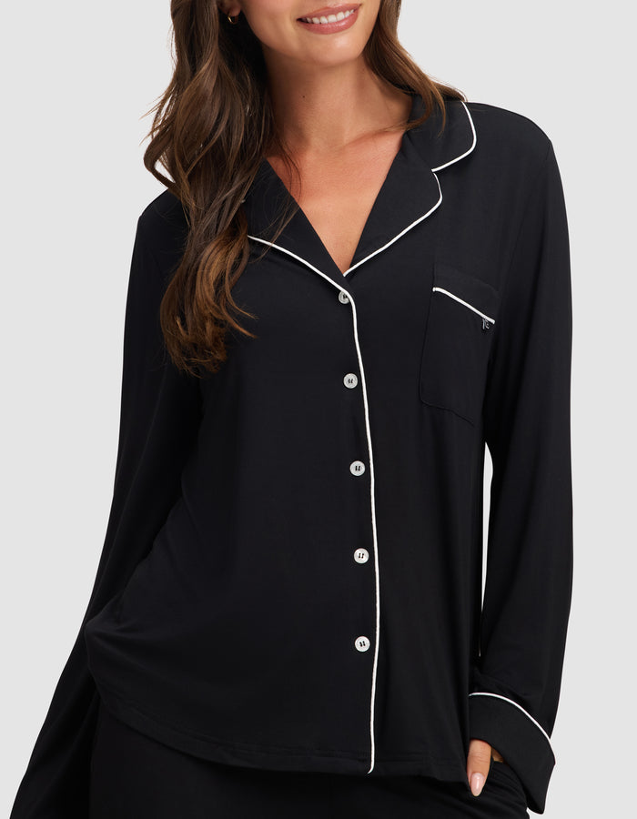 A woman smiles with one hand in her pocket, wearing the HIDE Women's Bamboo Stretch-Knit Long Sleeve Pajama Top in black with white piping, white buttons, and a chest pocket against a plain background. |Color:Black