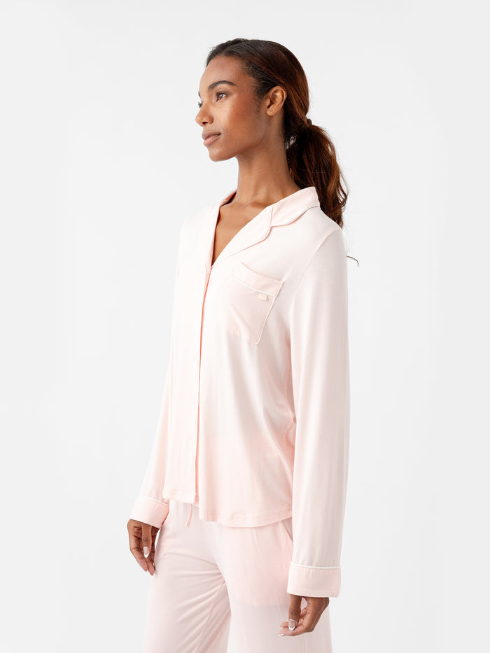 A person with long hair is wearing the Women's Long Sleeve Bamboo Pajama Top in Stretch-Knit TALL by HIDE, featuring a button-up shirt and pants in light pink. They are standing against a plain white background, looking off to the side with a relaxed expression. |Color:Blossom