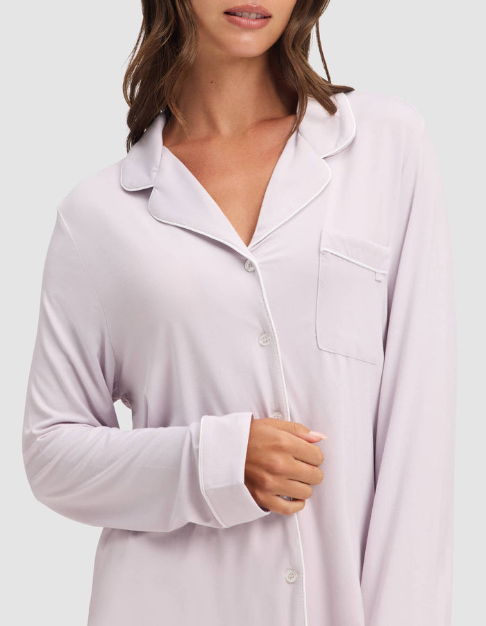 A woman wears the Cozy Earth Women's Bamboo Stretch-Knit Long Sleeve Pajama Top in light lavender with white piping, buttons, and a chest pocket. Her face is partially visible as she holds her right sleeve’s edge.|Color:Lilac