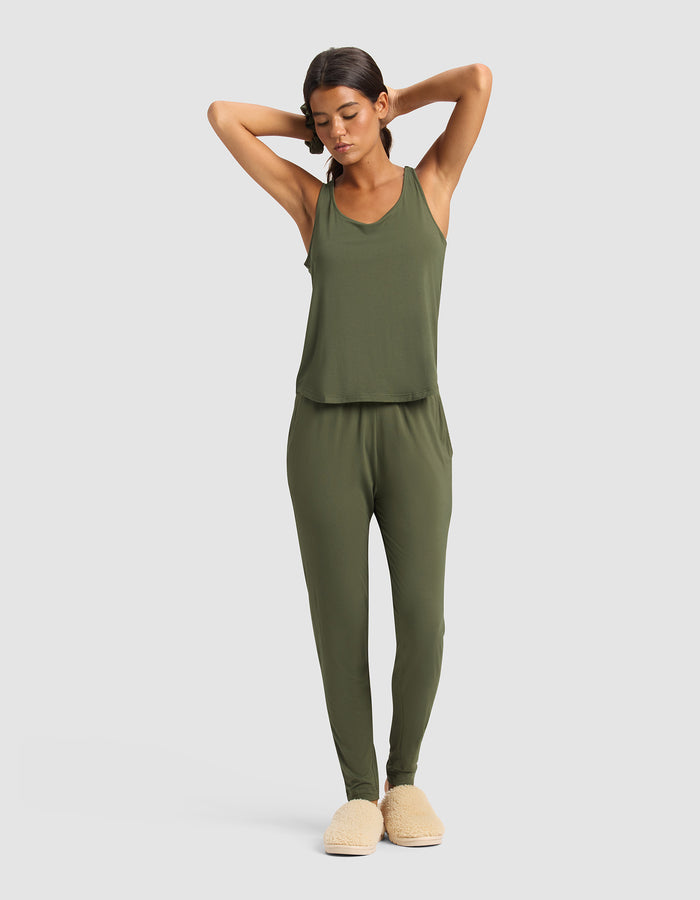 A woman stands with arms raised wearing the Cozy Earth Women's Bamboo Stretch-Knit Lounge Tank in olive green, matching jogger pants, and beige slippers against a plain light gray background.|Color:Olive