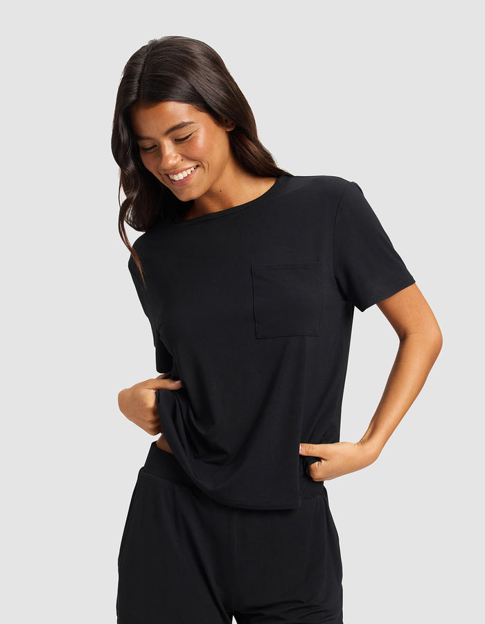 A woman with long dark hair smiles and looks down while wearing the Cozy Earth Women's Bamboo Stretch-Knit Lounge Tee & Pant Set in black against a plain light gray background.|Color:Black