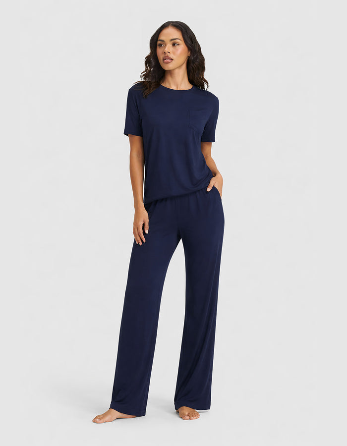 A woman stands barefoot in the Cozy Earth Women's Bamboo Stretch-Knit Lounge Tee & Pant Set—a matching short-sleeve top and wide-leg pant—posing against a plain light gray background.|Color:Navy