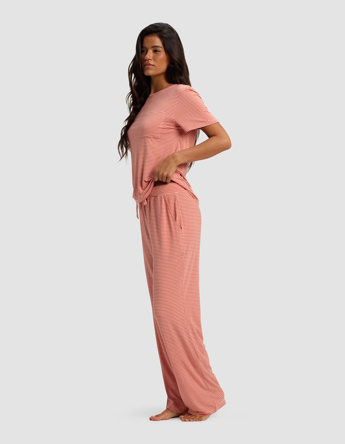 A woman stands barefoot in side profile, wearing Cozy Earth's Women's Bamboo Stretch-Knit Lounge Tee & Pant Set—loose-fitting with short sleeves and wide-leg pants—against a plain light gray background.|Color:Sunset Mini Stripe