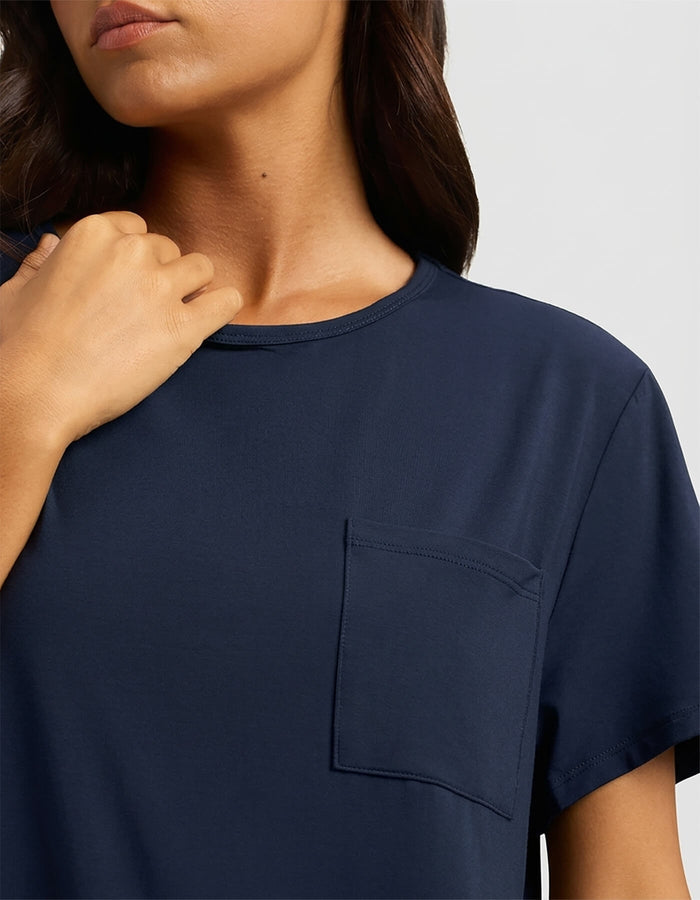 A woman with long brown hair touches her collarbone while wearing the Cozy Earth Women's Bamboo Stretch-Knit Lounge Tee, a navy blue short-sleeve shirt with a chest pocket, shown from shoulders to mid-torso.|Color:Navy