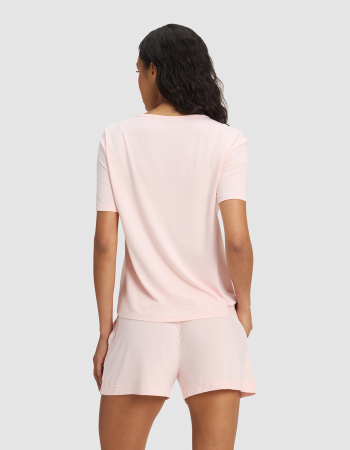 A woman with wavy dark hair is seen from the back, modeling the Cozy Earth Women's Bamboo Stretch-Knit Lounge Tee in light pink, paired with matching shorts against a plain light gray background.|Color:Blossom