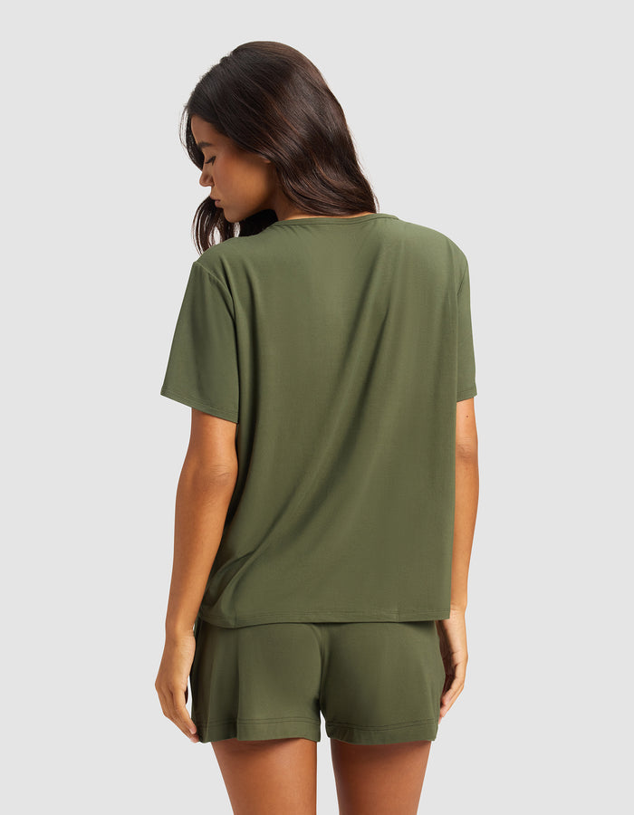 A woman with long brown hair faces away, wearing Cozy Earth's Women's Bamboo Stretch-Knit Lounge Tee in olive green, paired with matching shorts, set against a plain light gray background.|Color:Olive