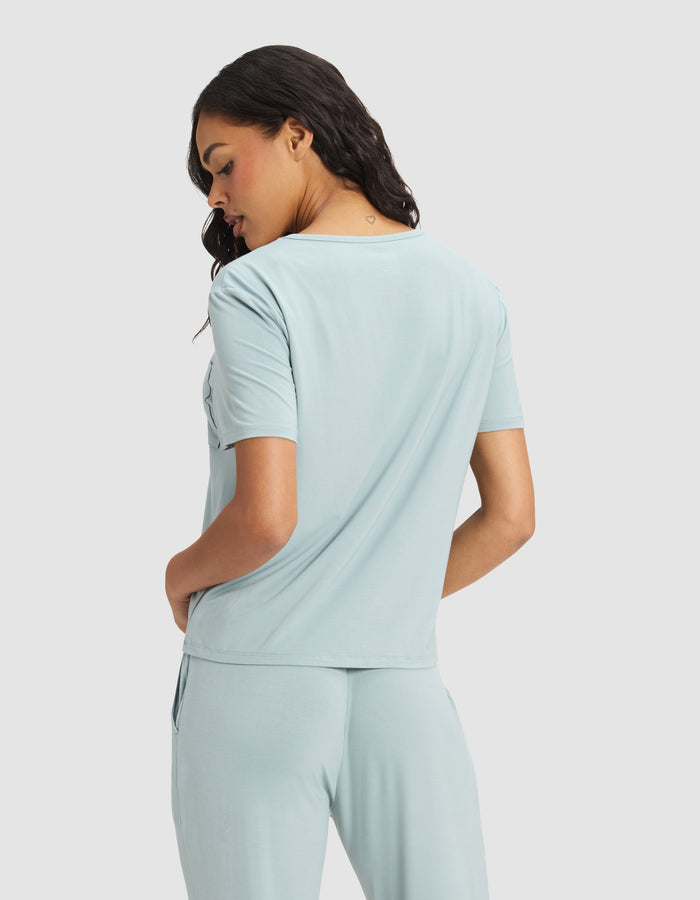 A woman faces away in a Cozy Earth Women's Bamboo Stretch-Knit Lounge Tee, light blue with short sleeves and matching pants. She turns her head slightly, hands in pockets, standing against a plain light background.|Color:Brook