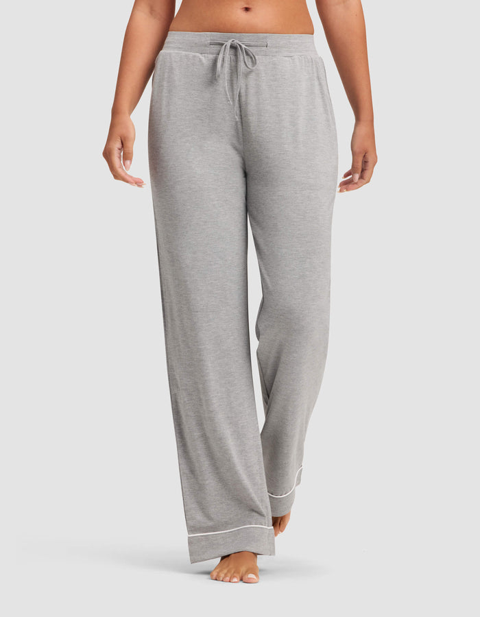 A woman wears Cozy Earth Women's Bamboo Stretch-Knit Pajama Pants in light gray, featuring a drawstring waist and white trim at the cuffs, standing barefoot against a plain background.|Color:Grey