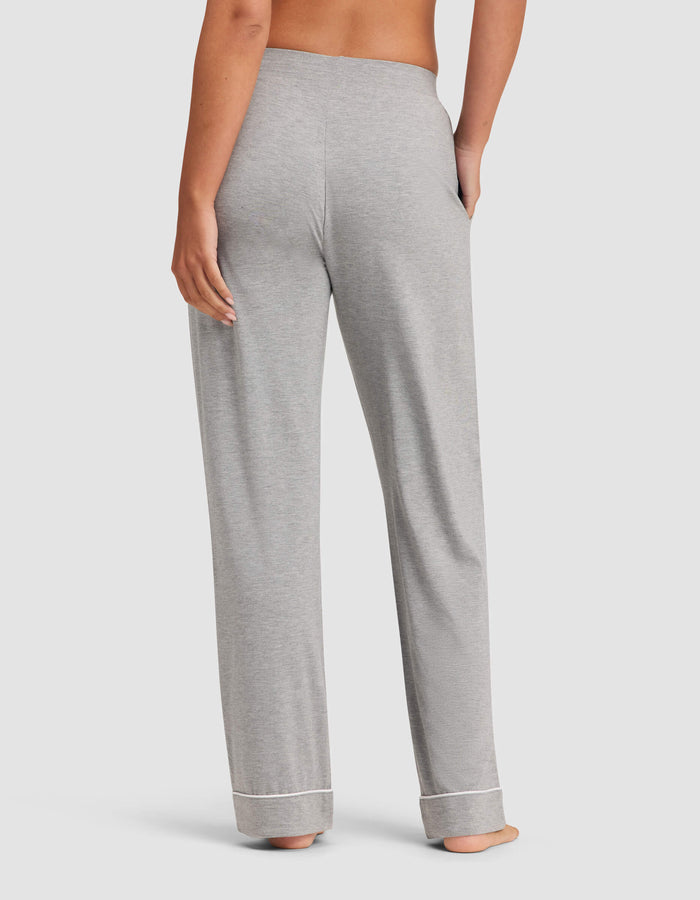A woman stands barefoot facing away, wearing Cozy Earth Women's Bamboo Stretch-Knit Pajama Pant in light gray with cuffed hems and one hand in a pocket, against a plain light background.|Color:Grey