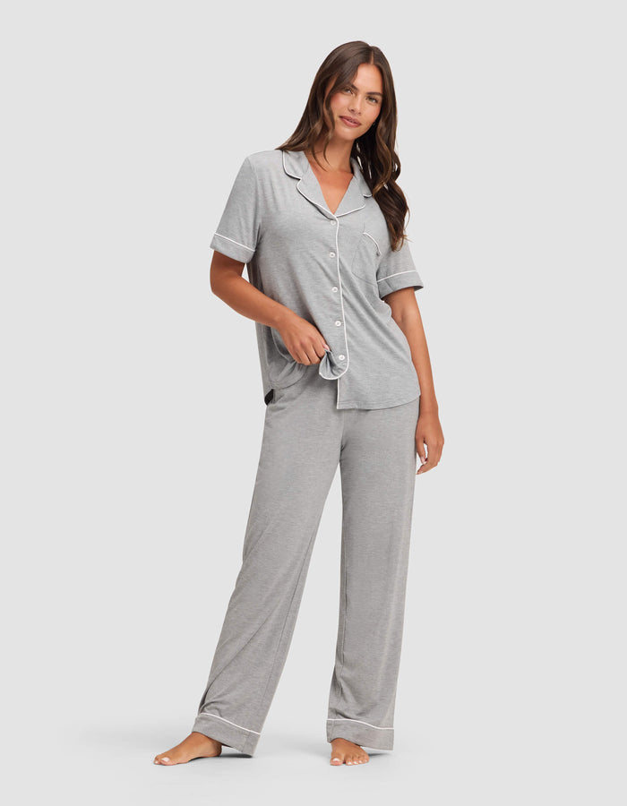 A woman with long brown hair stands barefoot against a plain backdrop, wearing Cozy Earth’s Women's Bamboo Stretch-Knit Pajama Pant in light gray, paired with a matching short-sleeve, collared top with white trim and buttons.|Color:Grey