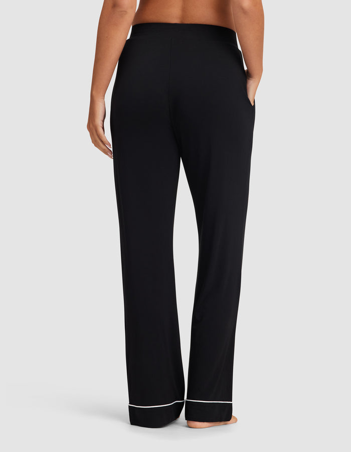 A woman stands barefoot, shown from the back, wearing Cozy Earth Women's Bamboo Stretch-Knit Pajama Pant in black with pockets and white hem trim against a plain light background.|Color:Black
