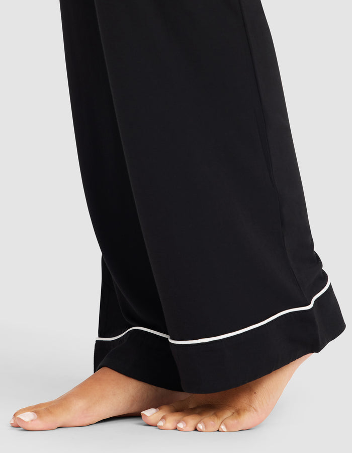 A close-up of bare feet on the floor in HIDE Women's Bamboo Stretch-Knit Pajama Pant, featuring black fabric with white piping on the hem, set against a plain light background.|Color:Black