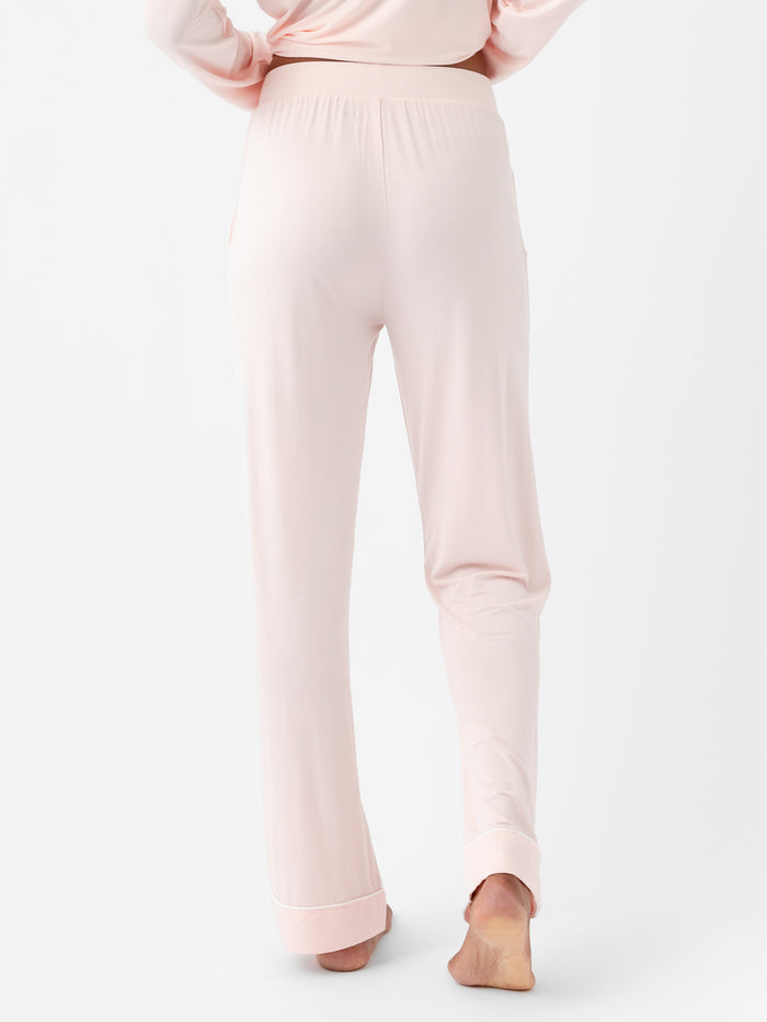 A person wearing blush pink Women's Stretch-Knit Bamboo Pajama Pants TALL by HIDE, shown from the back. The soft fabric pants feature an elastic waistband and gently drape over their legs. The person is barefoot, standing on their left foot with their right foot slightly lifted against a white background. |Color:Blossom