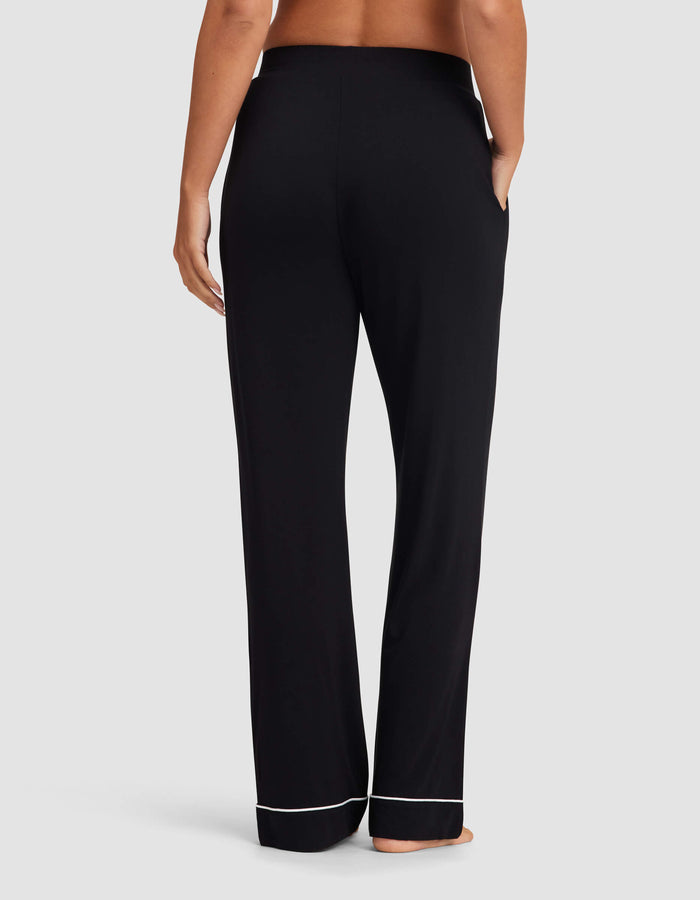 A woman stands barefoot facing away, wearing Cozy Earth Women's Bamboo Stretch-Knit Pajama Pant in black with white piping at the cuffs and hands in pockets against a plain light gray background.|Color:Black