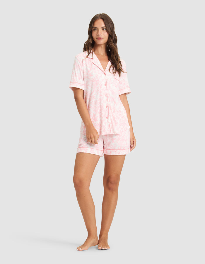A woman stands barefoot wearing HIDE's Women's Bamboo Stretch-Knit Pajama Shorts and a matching light pink, short-sleeve button-up top with a subtle pattern, set against a plain light gray background. |Color:Tulip Toile