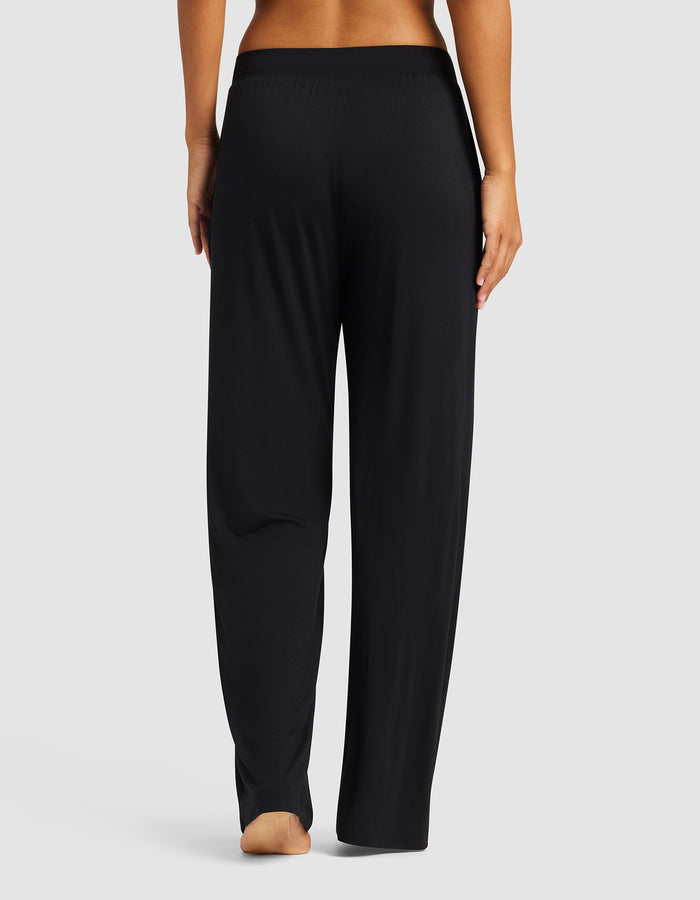 A person stands barefoot, facing away, wearing Cozy Earth’s Women's Bamboo Stretch-Knit Pant in black with an elastic waistband, set against a plain light gray background.|Color:Black