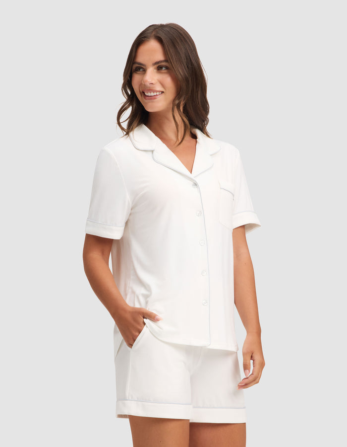 A woman smiles with her hands in her pockets, modeling the Cozy Earth Women's Bamboo Stretch-Knit Short Sleeve Pajama Set—a white button-up top and shorts—against a plain light gray background. |Color:Ivory