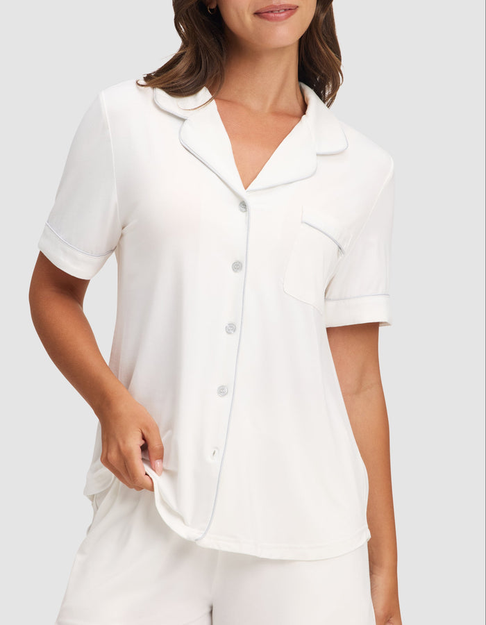 A woman wears the HIDE Women's Bamboo Stretch-Knit Short Sleeve Pajama Top with buttons and a chest pocket, paired with matching white shorts, standing against a plain light background. |Color:Ivory