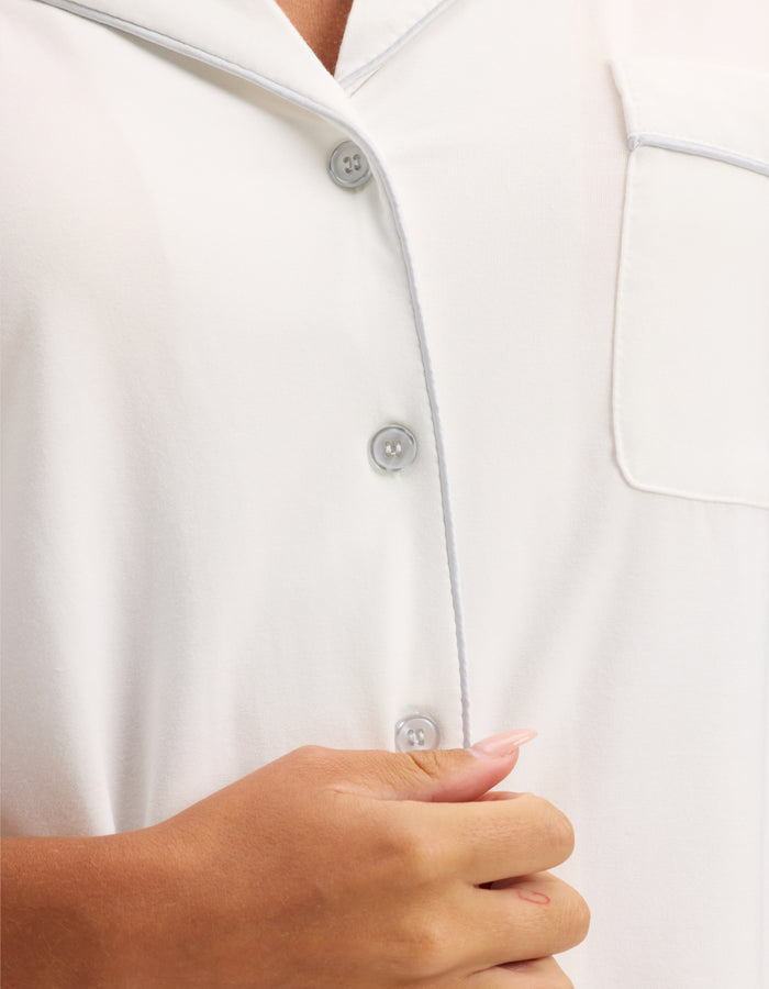 A close-up of a person wearing HIDE's Women's Bamboo Stretch-Knit Short Sleeve Pajama Top, holding the fabric near the buttons with their left hand, showing pale pink manicured nails. |Color:Ivory