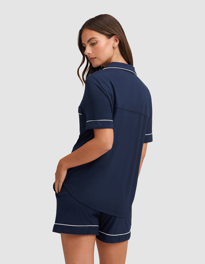A woman wearing the HIDE Women's Bamboo Stretch-Knit Short Sleeve Pajama Top in navy blue with white piping faces away, glancing over her shoulder with a hand in her shorts pocket against a plain light background. |Color:Navy