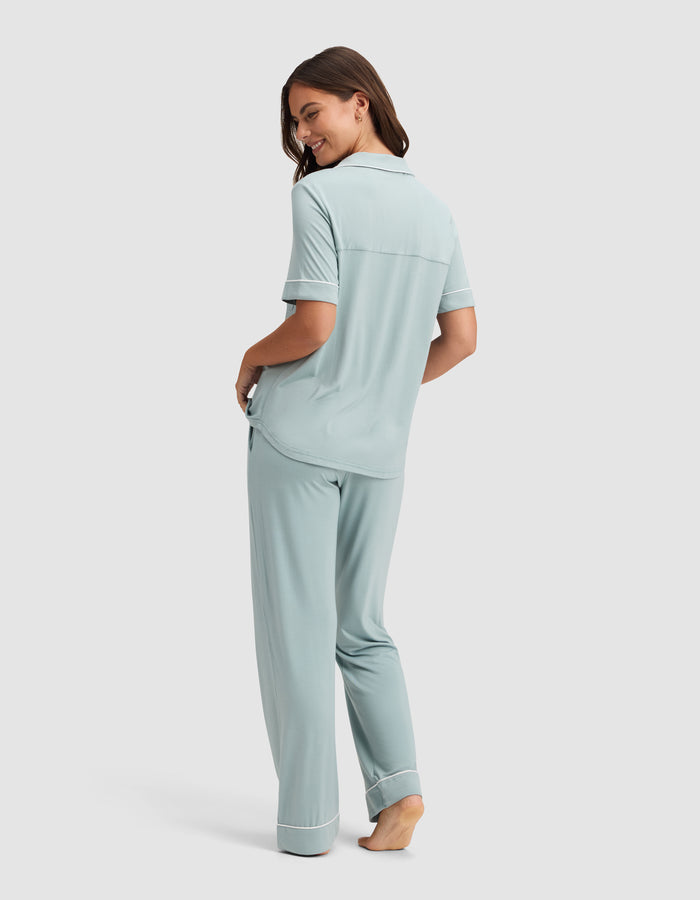 A woman, barefoot and smiling over her shoulder, models the Cozy Earth Women's Bamboo Stretch-Knit Short Sleeve & Pant Pajama Set in light blue with white trim against a simple background. |Color:Brook