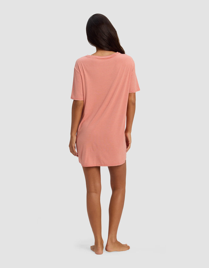 A woman stands barefoot facing away, wearing the Cozy Earth Women's Bamboo Stretch Knit Sleep Dress in pink and white stripes. The loose, short-sleeved dress falls above her knees. Her long dark hair is down against a light gray background.|Color:Sunset Mini Stripe