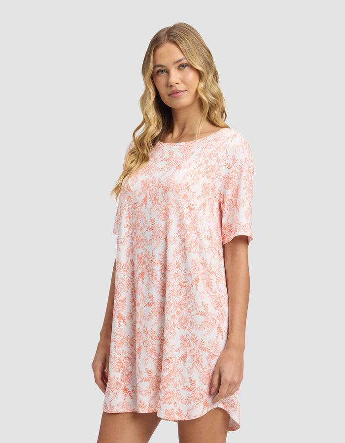 A woman with long blonde hair models the Cozy Earth Women's Bamboo Stretch-Knit Sleep Dress, a loose, short-sleeved light pink dress with an orange floral pattern, against a plain light gray background.|Color:Melon Toile