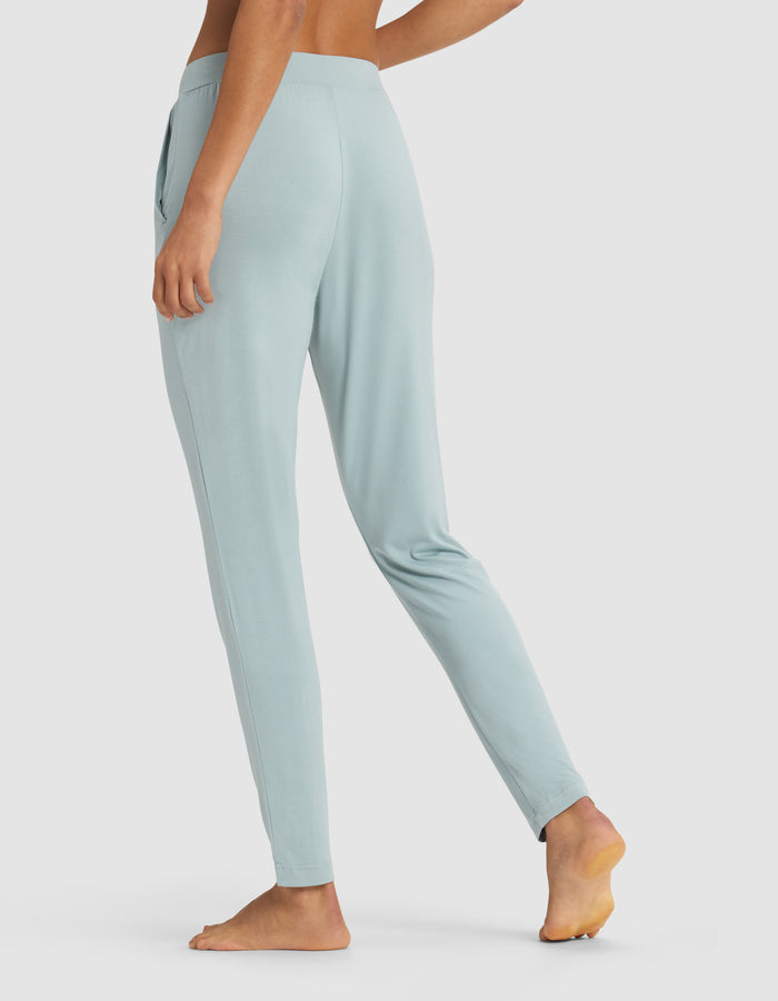 A person stands barefoot on tiptoe, wearing Cozy Earth’s Women’s Bamboo Stretch-Knit Tapered Pant in light blue, shown from the back and side. The high-waisted, slim-fit jogger is displayed against a plain background with the image cropped from the upper chest down.|Color:Brook