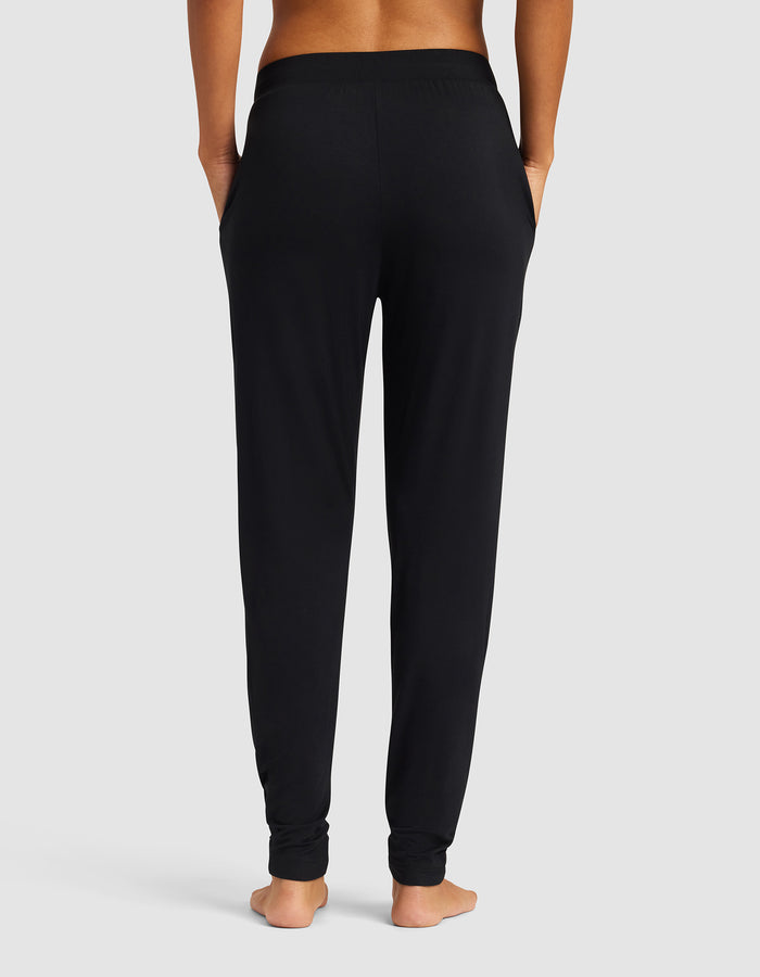 A woman stands barefoot facing away from the camera, wearing Cozy Earth's Women's Bamboo Stretch-Knit Tapered Pant in black with hands in pockets against a plain light gray background.|Color:Black