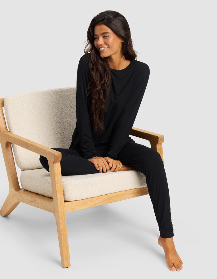 A woman with long dark hair sits barefoot on a light wooden chair, smiling and looking to the side while wearing Cozy Earth Women's Bamboo Stretch-Knit Tapered Pants and a matching black long-sleeve top.|Color:Black