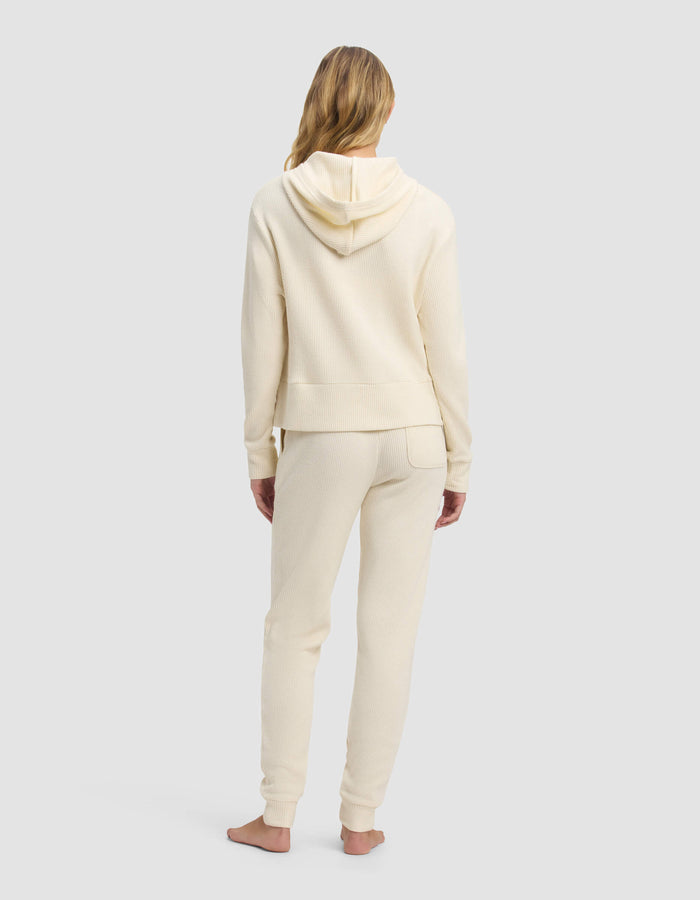 A woman with long blonde hair stands barefoot, facing away, wearing the Cozy Earth Women's Blake Waffle Hoodie & Jogger Set in cream against a plain light gray background.|Color:Coconut