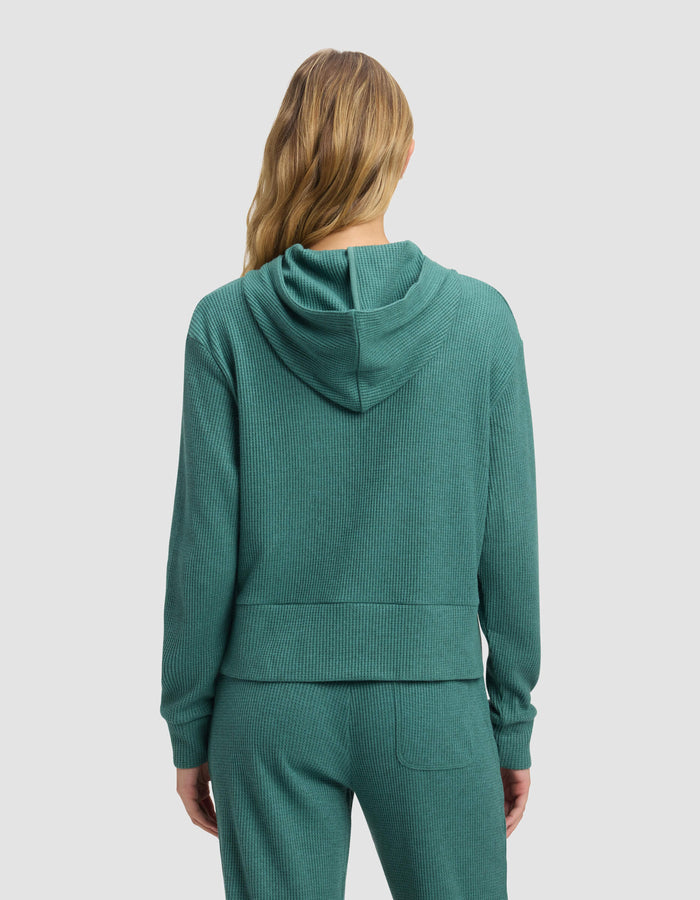 A woman with long, wavy, blonde hair is seen from the back wearing Cozy Earth's Women's Blake Waffle Hoodie in teal with matching pants against a plain, light background.|Color:Sea Green
