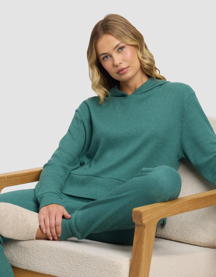 A blonde woman sits relaxed on a beige armchair, wearing the Cozy Earth Women's Blake Waffle Hoodie in teal, with one arm resting on the chair and her legs crossed.|Color:Sea Green