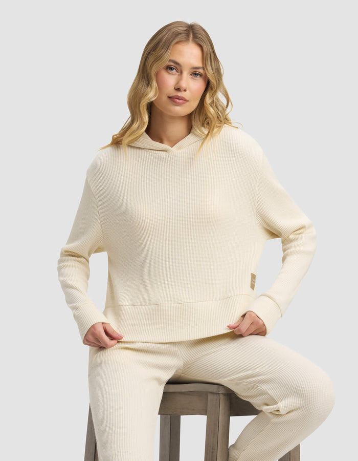 A woman with long, wavy blonde hair wears the Cozy Earth Women's Blake Waffle Hoodie in cream, paired with matching pants, while sitting on a wooden stool against a plain light gray background.|Color:Coconut
