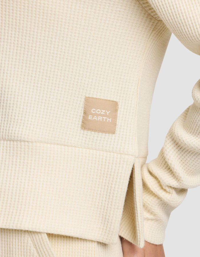 Close-up of the Women's Blake Waffle Hoodie by Cozy Earth in cream, showcasing its textured fabric and a stitched "COZY EARTH" tag. The wearer’s arm is bent at the elbow.|Color:Coconut