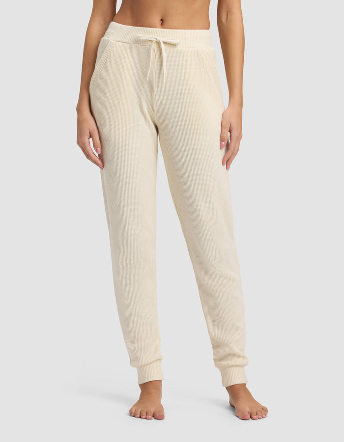 A person wears Cozy Earth's Women's Blake Waffle Jogger in cream, featuring a ribbed knit, drawstring waist, and pockets, while standing barefoot against a plain background.|Color:Coconut