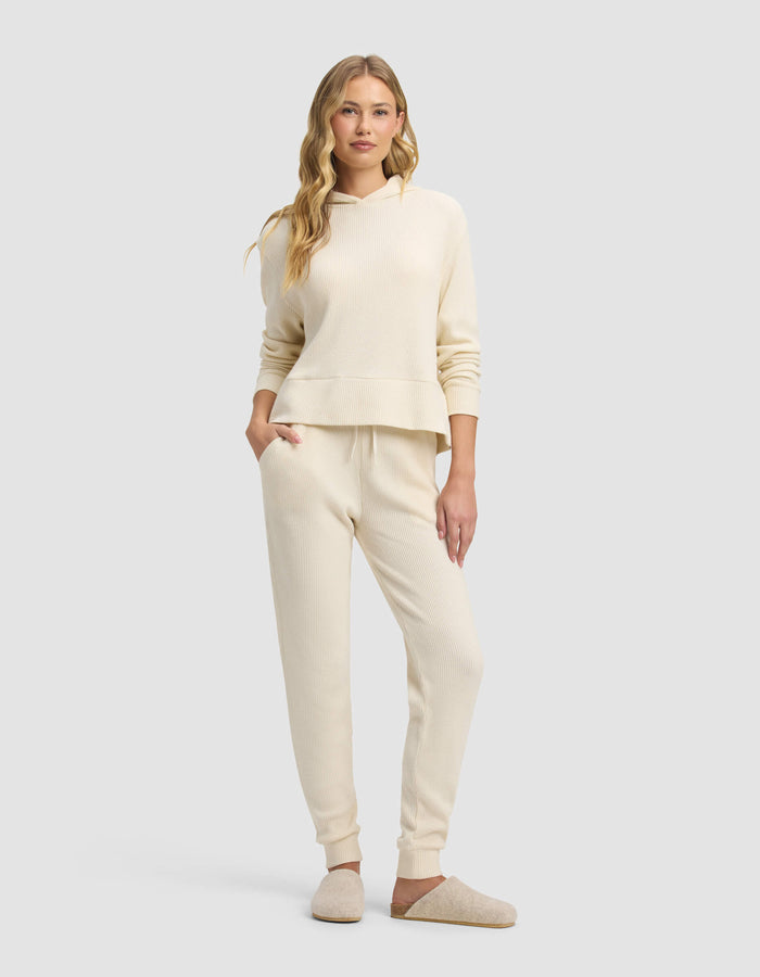 A woman with long wavy blonde hair stands against a plain background, wearing Cozy Earth's cream Women's Blake Waffle Jogger and matching hooded sweatshirt, paired with beige slippers. One hand rests in her pocket.|Color:Coconut