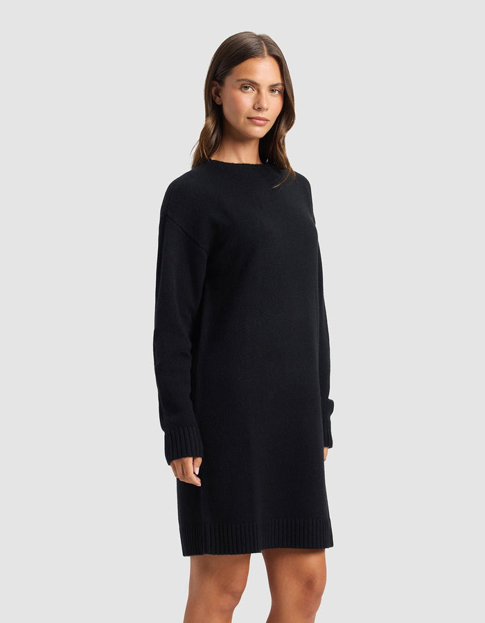 A woman with straight brown hair and a neutral expression stands sideways in a plain light gray setting, modeling the Cozy Earth Women's Brooklyn Sweater Dress—an elegant, long-sleeved, knee-length black dress.|Color:Jet Black