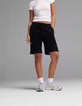 A person stands against a plain grey background wearing Cozy Earth Women’s Brushed Bamboo Bermuda Shorts, paired with a white fitted t-shirt, white socks, and white sneakers. Their hands are in their pockets and their face is not visible.|Color:Black