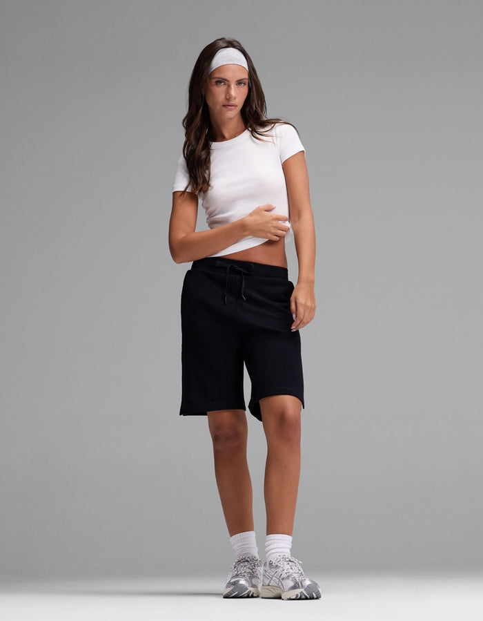 A person stands against a plain gray background wearing Cozy Earth’s Women’s Brushed Bamboo Bermuda Shorts, paired with a white headband, white crop top, white socks, and gray sneakers.|Color:Black