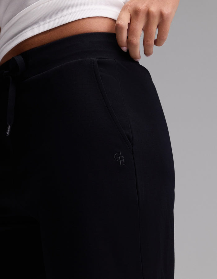 A person wearing Cozy Earth’s Women’s Brushed Bamboo Bermuda Shorts in black and a white top, with one hand resting on their hip. The shorts feature an embroidered “G B” logo near the pocket.|Color:Black