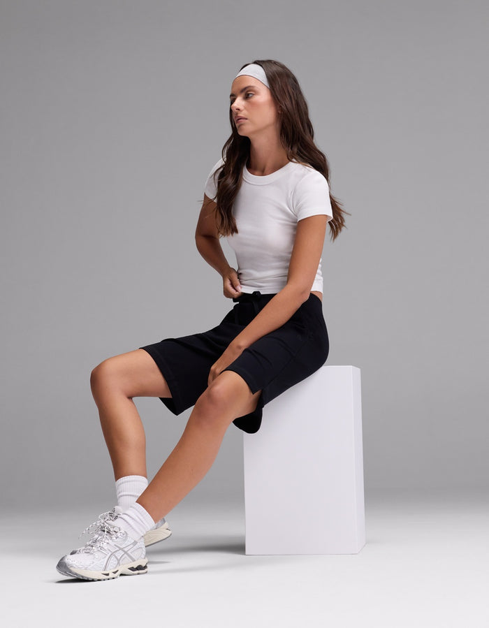 A woman wearing Cozy Earth’s Women’s Brushed Bamboo Bermuda Shorts, paired with a white t-shirt, white sneakers, and a gray headband, sits on a white block against a plain gray background, looking to the side.|Color:Black