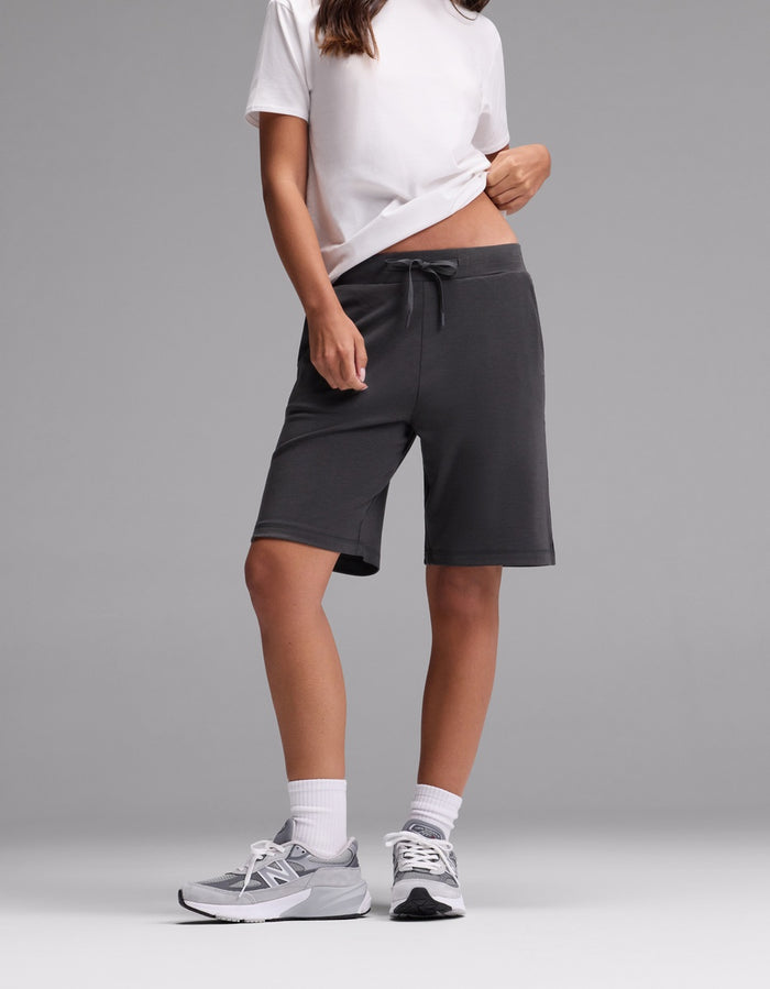 A person wearing a white t-shirt, Cozy Earth Women’s Brushed Bamboo Bermuda Shorts in dark gray, white socks, and gray sneakers stands against a plain gray background. Their face is not visible.|Color:Charcoal