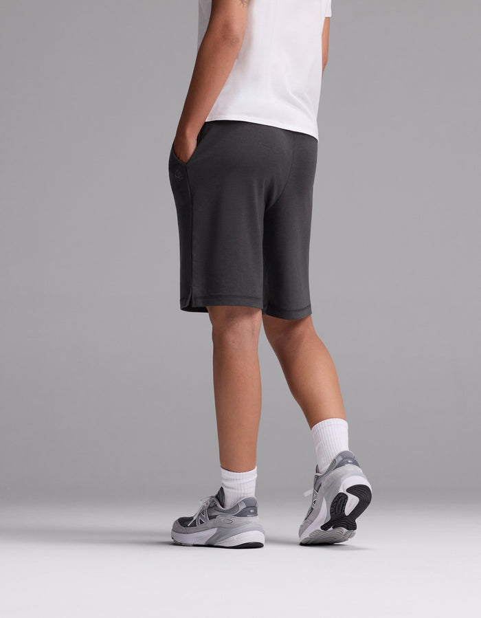 A person models Cozy Earth’s Women’s Brushed Bamboo Bermuda Shorts in gray, styled with a white t-shirt, white socks, and gray sneakers. They stand facing away against a plain gray background with one hand in their pocket.|Color:Charcoal