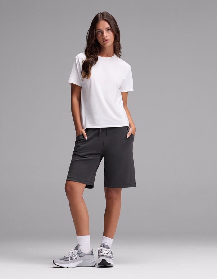 A woman stands against a gray background wearing Cozy Earth’s Women’s Brushed Bamboo Bermuda Shorts in dark gray, paired with a white t-shirt, white socks, and gray sneakers. Her hands are in her pockets as she looks slightly to the side.|Color:Charcoal