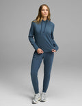 A woman with long blonde hair stands on a gray backdrop wearing the Cozy Earth Women’s Brushed Bamboo Hoodie & Jogger Set in blue, paired with white running shoes. She is holding part of her sweatshirt.|Color:Dawn