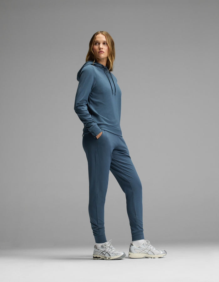 A woman stands sideways with her hands in the pockets of the Cozy Earth Women’s Brushed Bamboo Hoodie & Jogger Set, paired with gray sneakers, against a plain gray background.|Color:Dawn