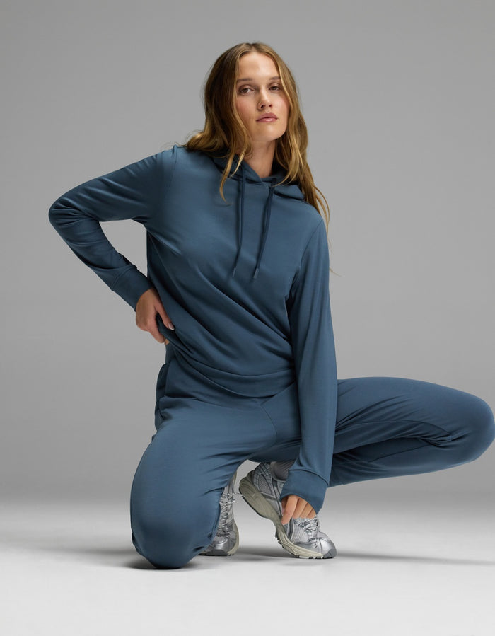 A woman showcases the Cozy Earth Women’s Brushed Bamboo Hoodie & Jogger Set in blue, kneeling on one knee with a hand on her hip against a plain gray background. She pairs the outfit with silver athletic shoes, and her long wavy hair cascades over her shoulders.|Color:Dawn