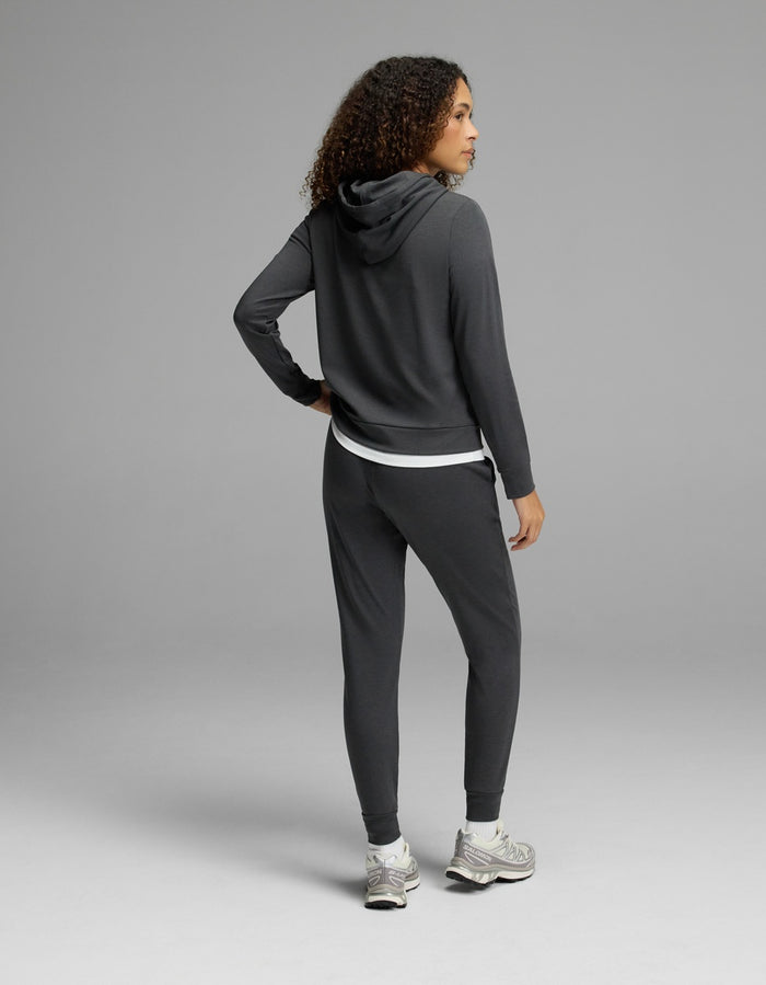 A woman with curly hair, dressed in the Cozy Earth Women’s Brushed Bamboo Hoodie & Jogger Set in a dark color, stands with one hand on her hip and faces away from the camera against a plain gray background.|Color:Charcoal